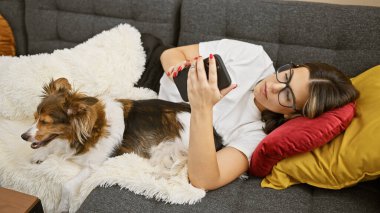 Genç bir İspanyol kadın, köpeğiyle birlikte kanepede uzanıyor, akıllı telefon kullanıyor, rahat bir ev ortamında..