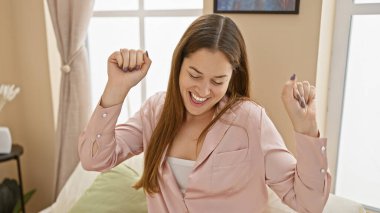 Uzun saçlı, gülümseyen kadın pembe gömlekli, parlak bir odada kutlama yapıyor.