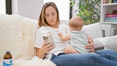 Rahat anne, kanepede oturuyor, akıllı telefonuyla mesajlaşırken ciddi bir ifade takınıyor, küçük kızıyla evde güzel bir anı paylaşıyor.