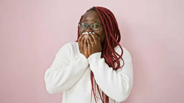 Adult african american woman with braids covering her mouth over a pink isolated background