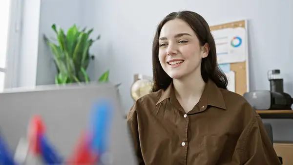 植物やチャートのぼやけた背景を持つ彼女のオフィスデスクで働くカジュアルな服装で笑顔の若い女性 — ストック写真