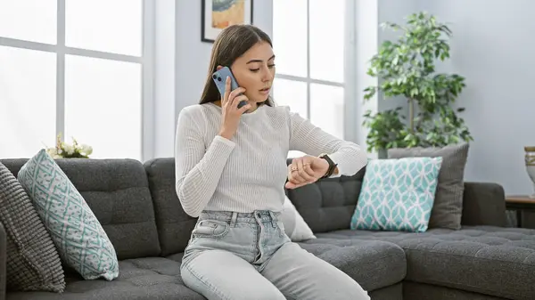 Hispanic woman talks on phone while checking time indoors, photo implies urgency and multitasking in a cozy living room.