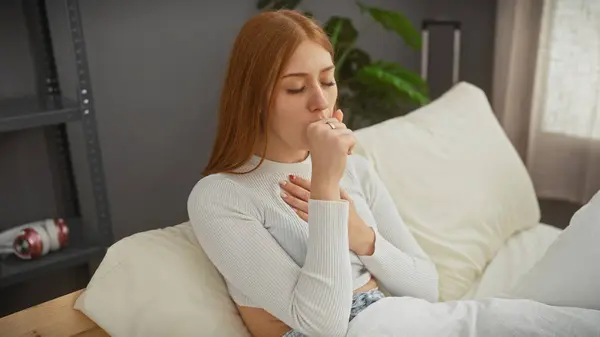A young caucasian woman with red hair is coughing while sitting on a bed indoors.