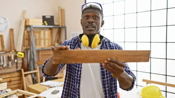 African american carpenter examines wood in a well-lit workshop wearing safety gear.
