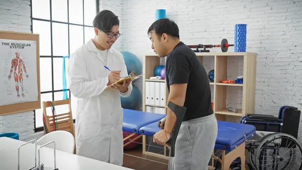 A male doctor with a clipboard talks to a man with a crutch in a well-equipped physiotherapy clinic interior