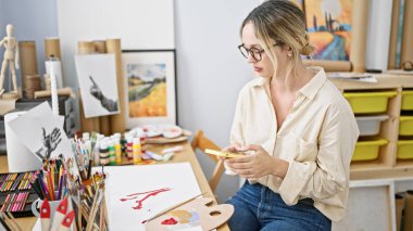 Sanat stüdyosunda akıllı telefon kullanan genç sarışın kadın sanatçı.