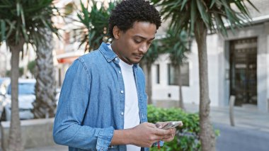 Kıvırcık saçlı yakışıklı genç adam Sunny City caddesinde akıllı telefon kullanıyor.