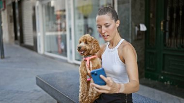 Köpekli beyaz bir kadın akıllı telefon kullanıyor. Caddedeki bankta oturuyor.