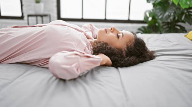 Kolay bir sabah, güzel bir İspanyol kadın pijamalarıyla yatakta uzanıyor, elleri başının üstünde, rahat bir yatak odasında kendini ifade ediyor.