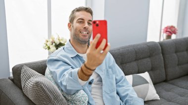 Yakışıklı, sakallı, modern bir oturma odasındaki kanepede selfie çeken bir İspanyol..