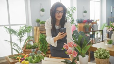 Orta yaşlı bir İspanyol kadın yeşillik dolu bir çiçekçinin içinde bitkilerin fotoğraflarını çekiyor..
