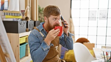 Önlüklü yakışıklı kızıl saçlı, kahve yudumlayan ve hareketli sanat stüdyosunda akıllı telefondan canlı sohbet eden genç bir sanatçı..