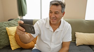 Gülümseyen orta yaşlı bir adam rahat oturma odasında akıllı telefon kullanıyor. Bağlantı ve rahatlama tasvir ediyor..