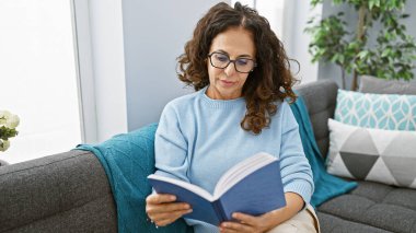Orta yaşlı, kıvırcık saçlı, rahat bir evin oturma odasında kitap okuyan ve sükuneti yansıtan bir İspanyol kadın..