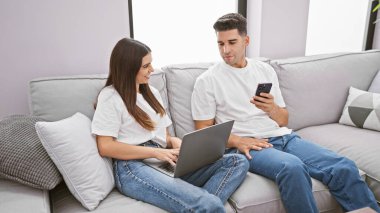 Gündelik giysiler içindeki bir kadın ve bir erkek rahat bir oturma odasındaki kanepede teknolojiyle meşgul olurlar.