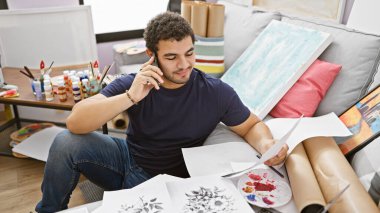 Genç bir adam, resimlerle ve sanat malzemeleriyle çevrili bir stüdyoda telefonla konuşurken sanat eserlerini eleştiriyor..