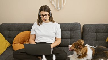 Konsantre olmuş genç bir İspanyol kadın köpeğiyle birlikte rahat bir oturma odasında dizüstü bilgisayarıyla çalışıyor..