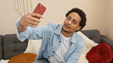 Kıvırcık saçlı yakışıklı bir adam rahat bir oturma odasında akıllı telefonuyla selfie çekiyor..