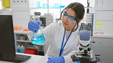 Laboratuvarda mavi kimyasalları analiz eden İspanyol kadın bilim adamı laboratuvar önlüğü ve güvenlik gözlüğü takıyor.