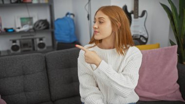 Beyaz saçlı kadın, rahat bir oturma odasında gri bir kanepede otururken işaret ediyor..