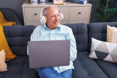 Middle age grey-haired man using laptop and headphones sitting on sofa at home