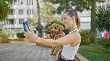 Köpeği gülümseyen beyaz bir kadın parkta akıllı telefonuyla selfie çekiyor.
