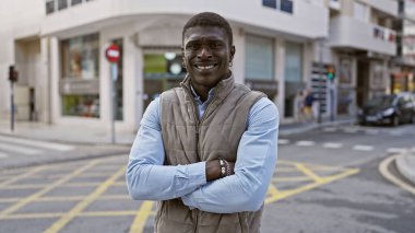 A confident african man with crossed arms smiles outside in an urban city environment. clipart