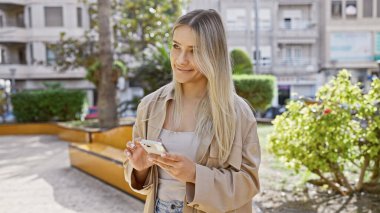 Çekici genç sarışın kadın telefonunu mutlu bir şekilde kullanıyor, dijital bir sohbete dalmış ve yeşil şehir parkında neşeli bir şekilde mesajlaşıyor.