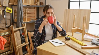 Genç bir İspanyol kadın ahşap işçiliği atölyesinde kahve molası veriyor. Etrafı alet ve ahşap parçalarıyla çevrili..