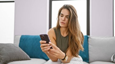 Genç ve güzel İspanyol kadın akıllı telefon kullanıyor. Kanepede oturuyor ve evde oturup düşünüyor.