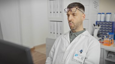 Gözlüklü ve sakallı İspanyol bir adam tıbbi laboratuvar odasında duruyor..
