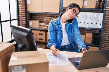 Young beautiful hispanic woman ecommerce business worker using laptop talking on smartphone at office