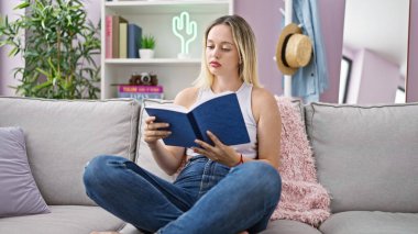 Young blonde woman reading book sitting on sofa at home