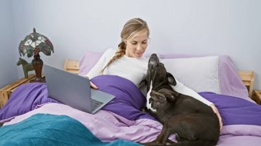 Genç beyaz kadın, laptopunu içeride labradoruyla yatakta kullanıyor.