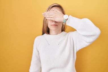 Sarı arka plan üzerine beyaz kazak giyen genç beyaz bir kadın. Gözleri elle kaplıyor. Ciddi ve üzgün görünüyor. Görme, saklama ve reddetme konsepti 