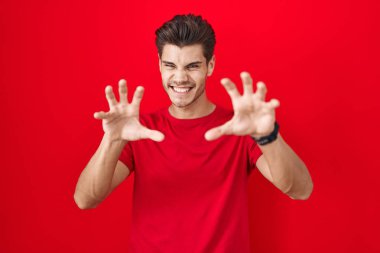 Kırmızı arka planda dikilen genç İspanyol adam komik bir şekilde pençe hareketi yapıyor, saldırgan ve seksi bir ifade. 