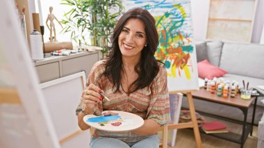 Parlak bir sanat stüdyosunda tuvale resim yaparken elinde palet tutan gülümseyen olgun bir kadın..