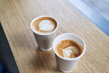 İki paket kapuçino, ahşap bir masada, kalp latte sanatı ile sıcak, kahve molası atmosferi taşır..