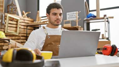 Sakallı genç adam marangozluk stüdyosunda dizüstü bilgisayarla çalışıyor, etrafı ahşap işçiliği aletleriyle çevrili..