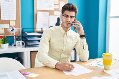 Young hispanic man business worker using smartphone writing on document at office