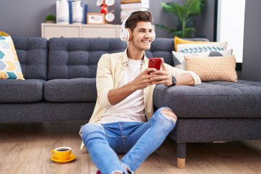 Young hispanic man listening to music sitting on floor at home