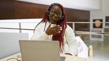 Ofis içi iş yerinde örgülü, kulaklık takan gülümseyen Afrikalı bir kadın.