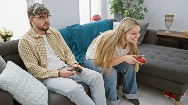 Oturma odasındaki gri bir kanepede oturan bir adam ve kadın bir video oyunu oynuyorlar.