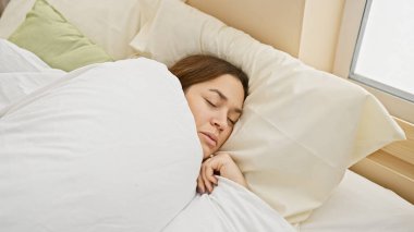 Huzurlu bir kadın aydınlık bir yatak odasında rahat bir şekilde uyuyor, sükuneti bembeyaz bir yatak ve sabah ışığı ile tasvir ediyor.