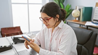 Modern ofis ortamında akıllı telefonuna odaklanmış profesyonel bir İspanyol kadın..