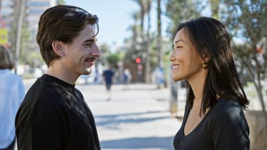 Güneşli bir şehir sokağında, gündelik kıyafetli bir kadın ve bir erkeği net bir şekilde gören gülümseyen ırklar arası bir çift..