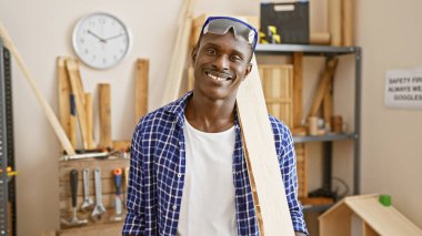 Bir marangoz atölyesinde güvenlik gözlüğü ve günlük giysiler takan gülümseyen Afrikalı bir adam..
