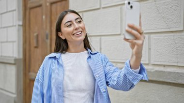 Latin kökenli genç bir kadın, çekici gülüşünü ve günlük tarzını sergileyerek şehir sokaklarında selfie çekiyor..