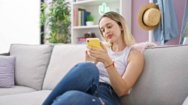 Evdeki kanepede oturan akıllı telefon kullanan genç sarışın kadın.