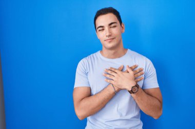 Mavi arka planda duran genç İspanyol adam elleri göğsünde, gözleri kapalı ve yüzünde minnet dolu bir jest ile gülümsüyor. sağlık kavramı. 